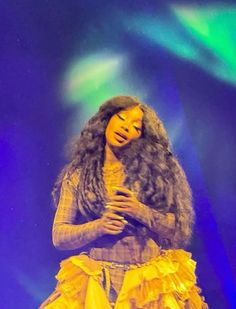 a woman with long hair standing in front of a stage wearing a yellow frock