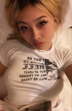 a woman laying in bed reading a book and looking at the camera while wearing a t - shirt that says, i may be to hell but at least all my friends will be there