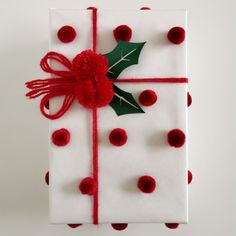 a present wrapped in white paper with red pom - poms and a holly on top