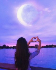 a girl is making a heart shape with her hands while looking at the moon over water