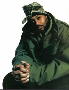 a man sitting down with his hands on his knees wearing a green jacket and hat