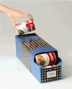 a hand is opening the top of a box with canned food in it on a white surface