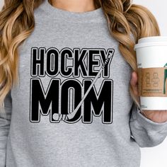 a woman holding a cup of coffee and wearing a hockey mom sweatshirt