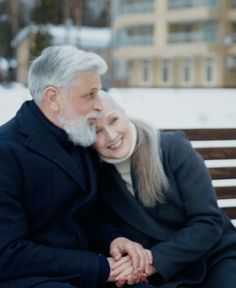 an older man and woman sitting on a bench in the snow smiling at each other