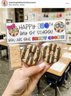 a person holding up two cookies in front of a sign that says happy 100th day of school you are one smart cookie