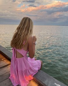 a woman in a pink dress sitting on a dock looking out at the water and clouds