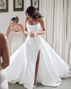 a woman in a white wedding dress is looking at herself in the mirror while another woman looks on