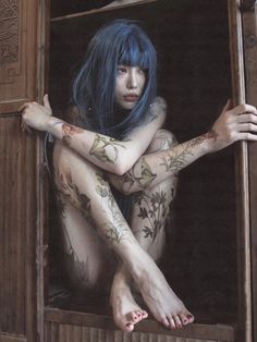 a woman with blue hair and tattoos on her body sitting in an open window sill