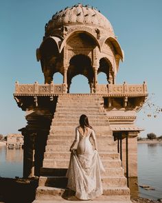 a woman in a white dress is standing on steps near a body of water and a gazebo