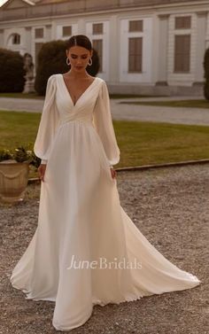 a woman standing in front of a large house wearing a white wedding dress with long sleeves