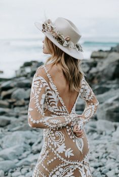 a woman in a dress and hat standing on rocks near the ocean with her back to the camera