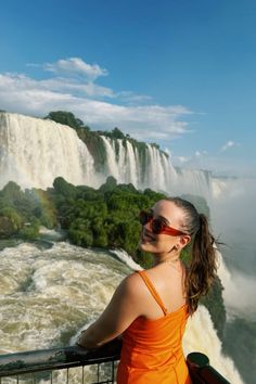 a woman in an orange dress standing at the edge of a waterfall looking into the distance