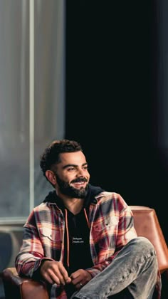 a man sitting on top of a brown chair in front of a black wall and smiling