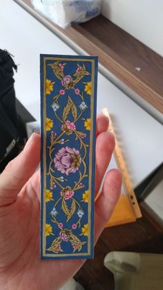 a hand holding up a piece of paper with flowers and vines on it, in front of a table