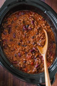 a slow cooker filled with chili and beans next to a wooden spoon on a table