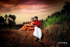 two people sitting on top of a dirt hill next to tall grass and palm trees