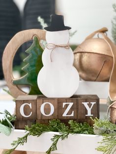 a wooden block that says cozy with a snowman on top and evergreen branches around it