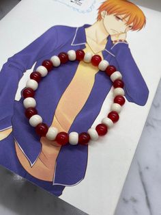 a bracelet with red and white beads sits on top of a piece of paper next to an anime character