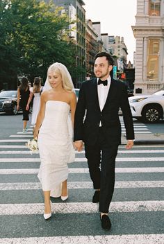 a bride and groom are walking across the street in their tuxedo attire, holding hands