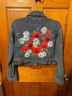 a denim jacket with red and white flowers on it hanging from a wooden closet door