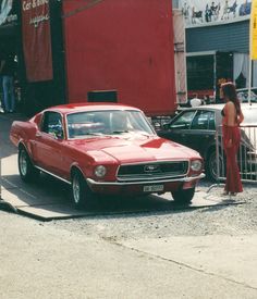 an old red car is parked in front of a woman standing next to the fence