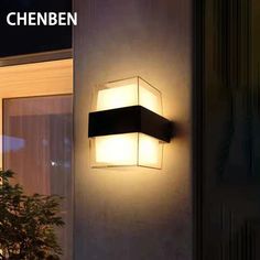 an outdoor wall light that is on the side of a building with plants in front of it