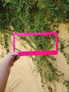 a hand holding up a pink license plate frame in front of a wall covered with green plants