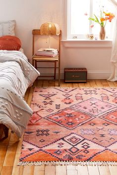 an orange and pink rug in a white room with a bed, window, and nightstand