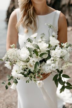 a woman in a white dress holding a bouquet of flowers and greenery by the water