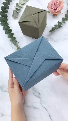 someone holding an origami box in front of some flowers and greenery on a marble surface