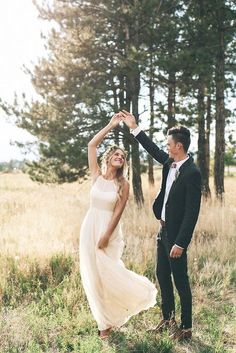 a young man and woman are dancing in the grass near some tall trees while holding each other's hands