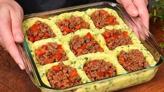 a person holding a casserole dish with meat and vegetables in it on a wooden table