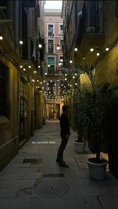 a man standing in an alley way with lights strung from the ceiling and potted plants on either side