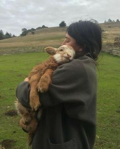 a woman holding a baby goat in her arms while standing on top of a lush green field