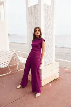 a woman leaning against a pillar in front of the ocean with her hands on her hips