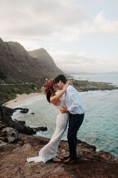 a bride and groom kissing on top of a cliff overlooking the ocean with mountains in the background