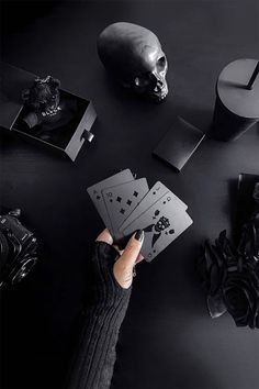 a person holding playing cards in their hands with skulls and other objects on the table