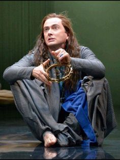 a man with long hair sitting on the floor in front of a green wall holding a gold ring