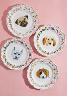 four plates with dogs painted on them sitting on a pink tableclothed surface in front of a pink wall