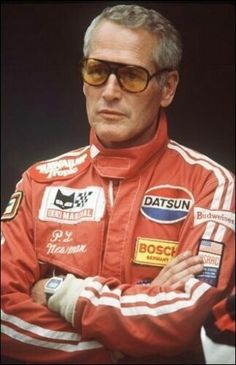 an older man in a red racing suit with his arms crossed, wearing sunglasses and looking off to the side