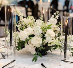 white flowers and greenery are arranged in glass vases on a table with silverware