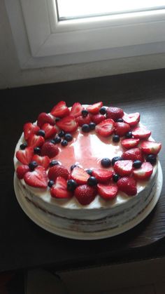 a white cake with strawberries and blueberries on top sitting on a table next to a window