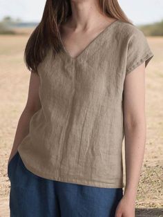 a woman standing in the middle of a field wearing a tan shirt and blue pants