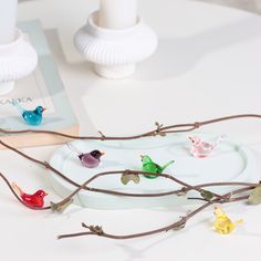 several small birds are sitting on branches in front of a book and vase with two candles