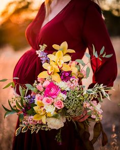 a pregnant woman in a red dress holding a bouquet of flowers and greenery with the sun setting behind her