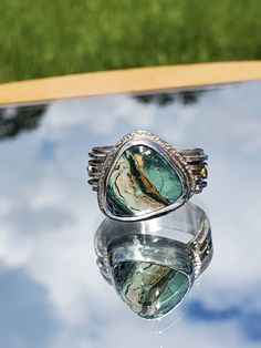 This ring features a 15 x 11mm Turquoise gemstone set in Sterling Silver and accented with two, 14kt gold balls.  The turquoise features bands of gold, cream, light green and dark green.  It is cut in a triangle shape and set at an angle to best compliment a woman's finger.  The band is 10 mm wide and features 4 individual bands of Sterling Silver to give it a multi-dimensional look.  The gold balls accent the gold in the turquoise. This is a one-of-a-kind ring that you will not see anywhere else. RETURN POLICY: I accept returns under the following conditions.  The jewelry must be returned in new condition.  Purchaser must contact me within 3 days of receipt of the jewelry.  The ring must be returned in the same jewelry box it was shipped in, and it must be returned vias USPS Priority Mail Elegant Green Chrysocolla Jewelry, Unique Turquoise Chrysoprase Jewelry, Bohemian Green Rings With Gemstone Accents, Unique Blue Emerald Jewelry, Unique Turquoise Ring With Gemstone Accents, Unique Green Turquoise Ring With Large Stone, Unique Turquoise Ring With Gemstone Accents As Gift, Green Turquoise Ring With Natural Stones, Unique Turquoise Chrysocolla Ring