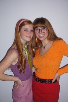 two women standing next to each other in front of a white wall and one is wearing an orange sweater