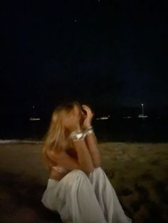 a woman is sitting on the beach at night with her hands behind her head and looking off into the distance