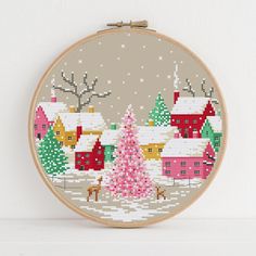 a cross stitch christmas scene with a pink tree in the middle and houses on the other side