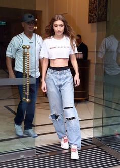 a woman in ripped jeans and a white crop top is walking out of a building